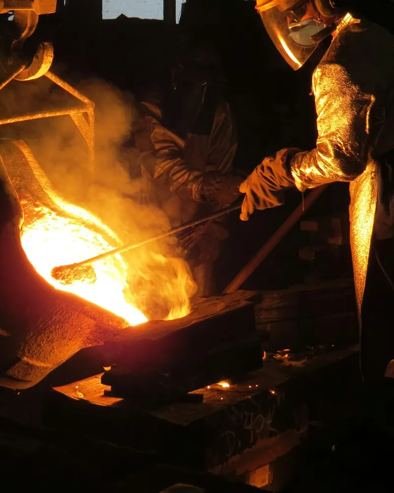 Fundidor com fato refletor e viseira a retirar a escória da superfície do metal fundido numa colher de vazamento